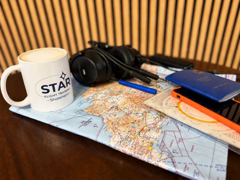 Flight planning materials, a headset and a pilot's licence arranged on a coffee table alongside a café latte in a branded mug, in the reception of STAR Flight Training, Shoreham.
